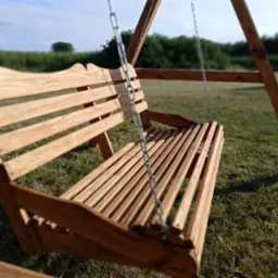 Drewniana huśtawka ogrodowa z oparciem o dekoracyjnym kształcie, zawieszona na łańcuchach. Tło to trawnik i zieleń, w oddali pole. Słoneczny dzień.