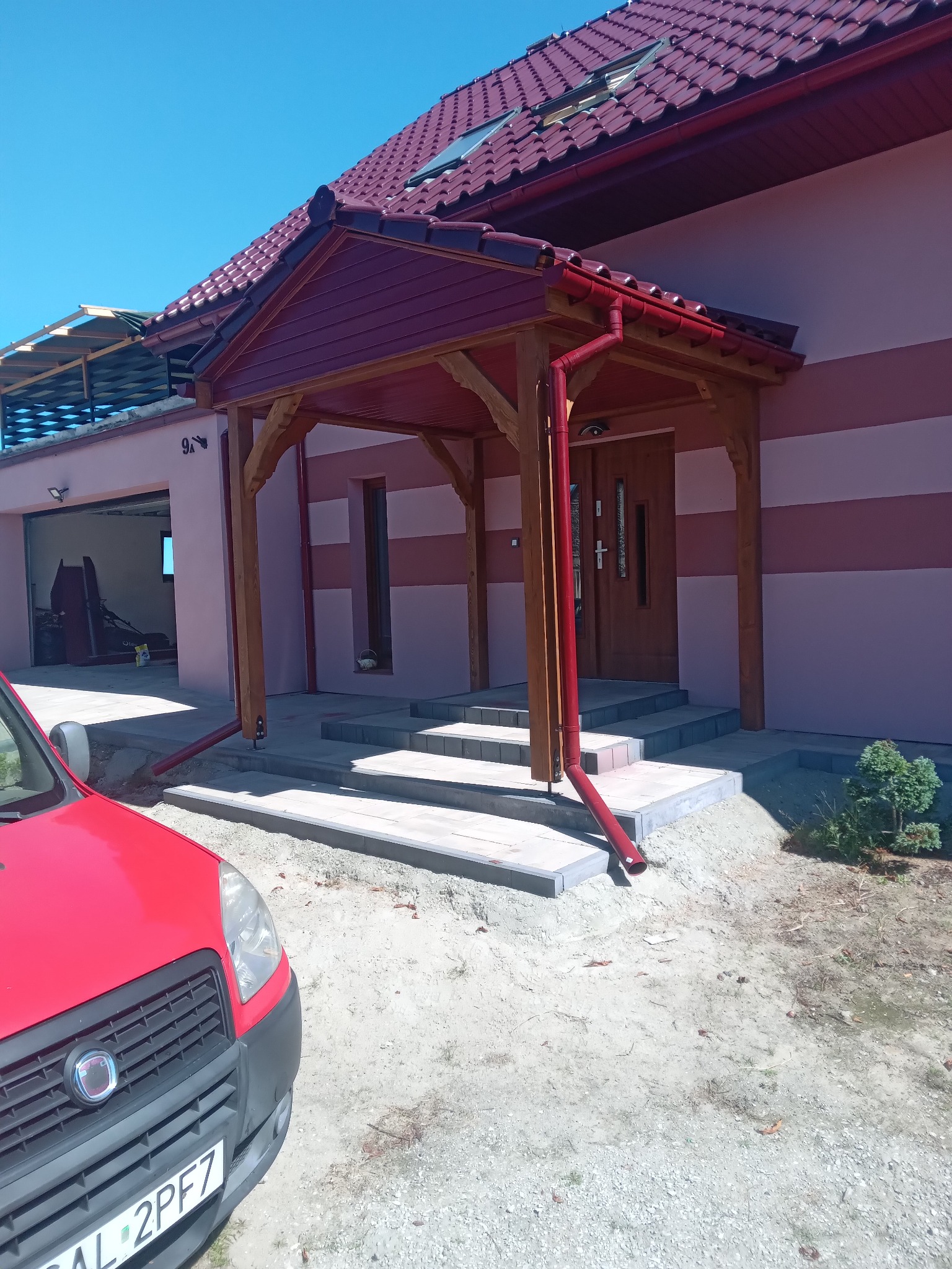 Zadaszenie wejścia do domu z drewnianą konstrukcją i czerwonymi rynnami. Widoczne schody wejściowe i fragment czerwonego samochodu.