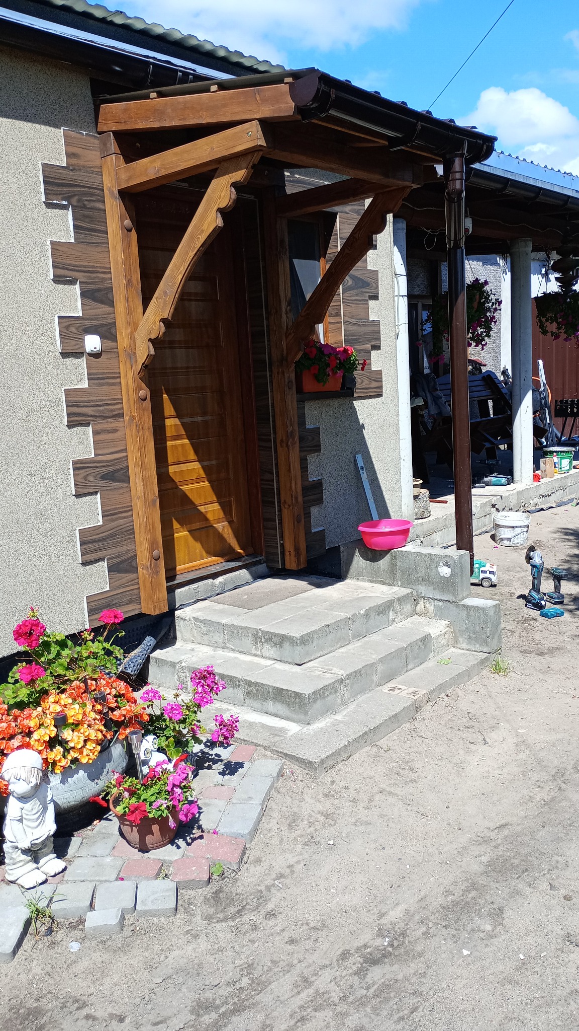 Drewniany daszek nad wejściem do domu, z doniczkami z kwiatami. Widoczne narzędzia i elementy wykończeniowe. Elementy dekoracyjne z boku schodów.