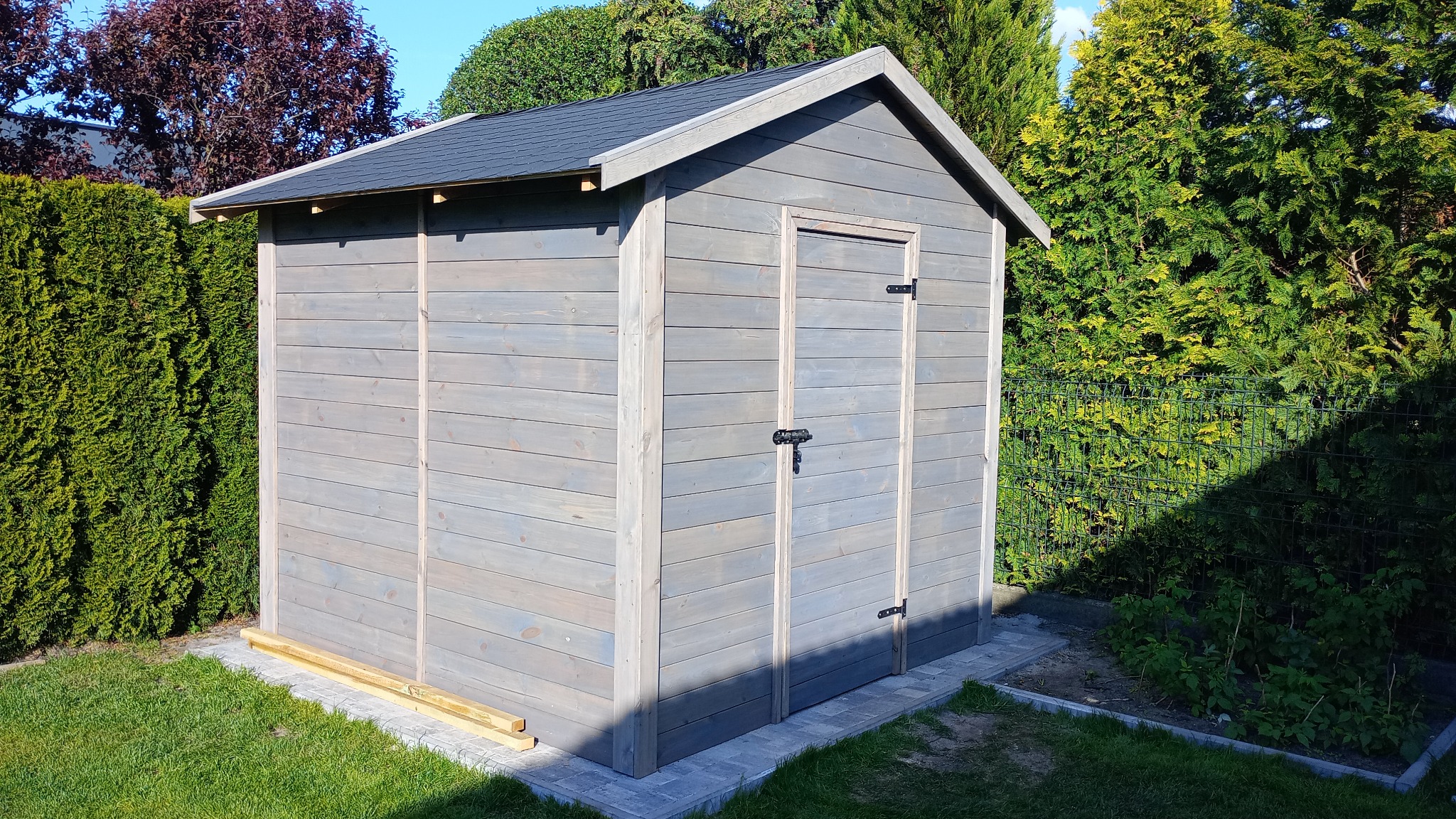 Szara, drewniana budka narzędziowa z czarnym dachem, umieszczona na kostce brukowej w ogrodzie. Widoczne krzewy i drzewa w tle.
