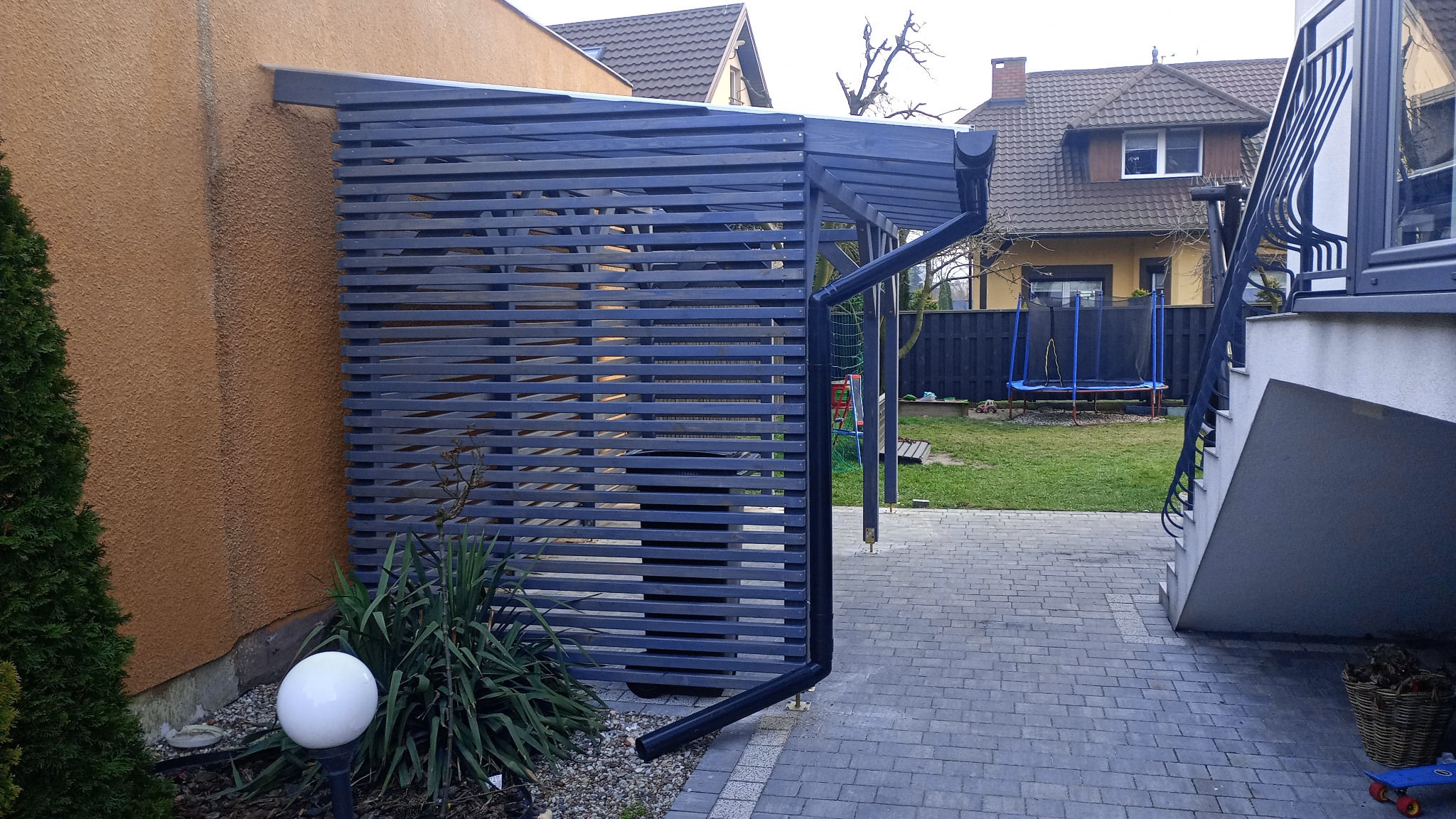 Ciemnoszara drewniana konstrukcja z poziomych listew, przylegająca do budynku. W tle widoczny ogród z trampoliną i dom.
