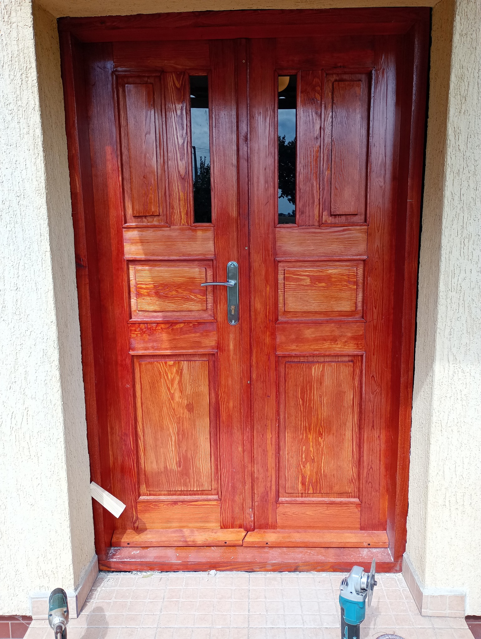 Lakierowane, dwuskrzydłowe drzwi wejściowe z szybkami, świeżo po renowacji w Aleksandrowie Kujawskim. Widoczne narzędzia pracy i niedoskonałości wykończenia.