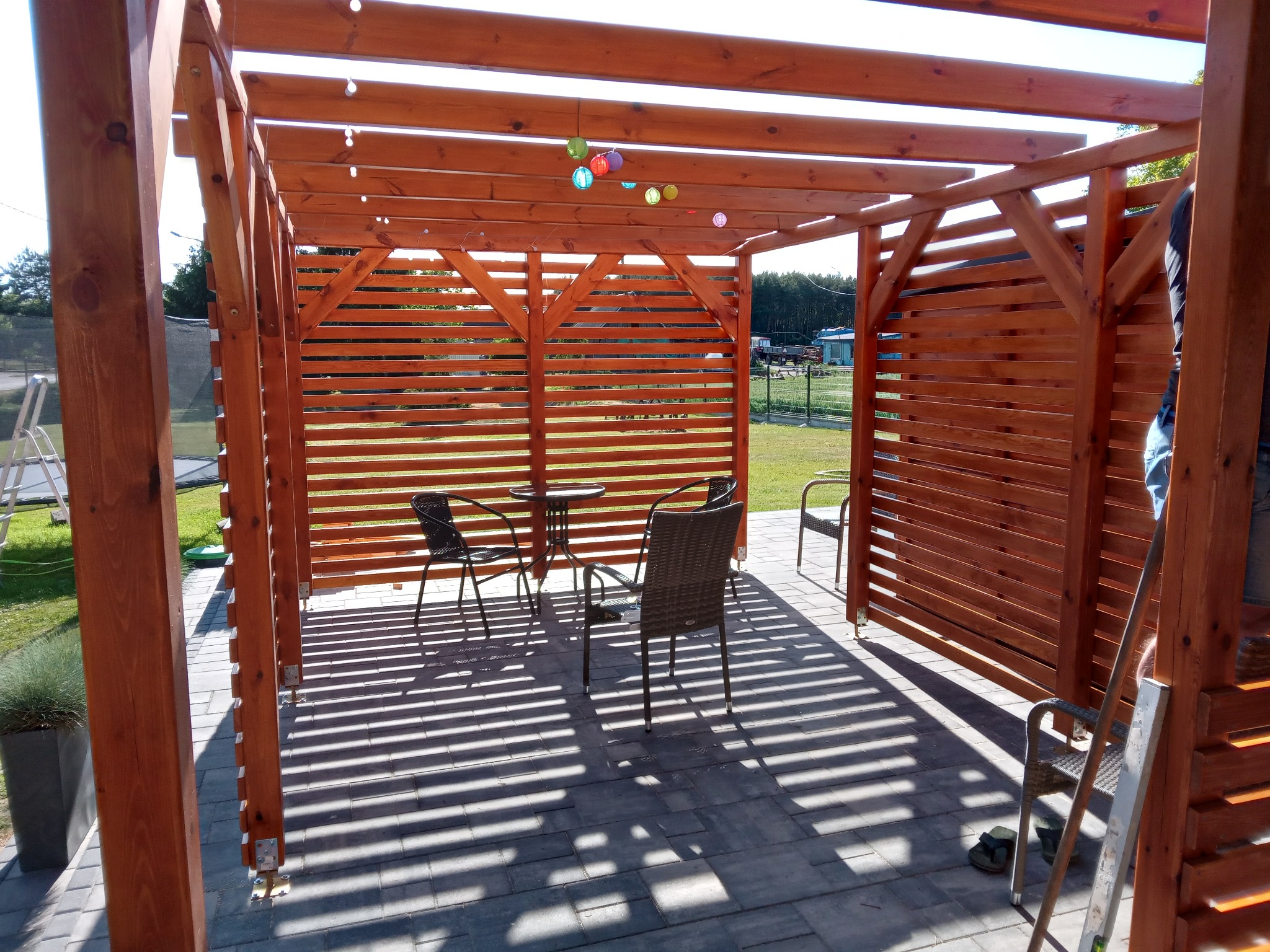 Drewniana pergola z dekoracyjnymi lampionami i meblami ogrodowymi na brukowanym tarasie, tworząca zacienioną strefę relaksu.