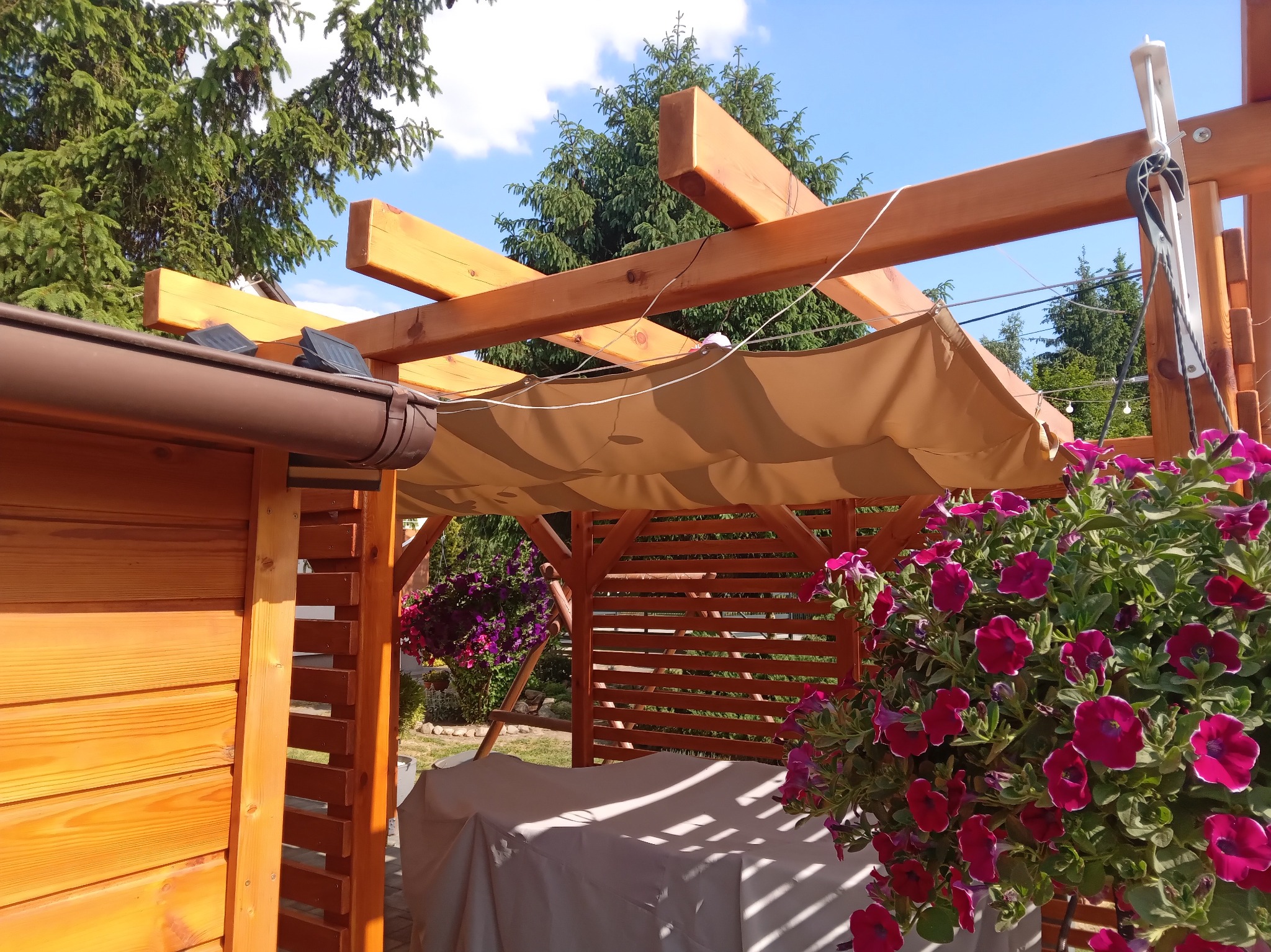 Drewniana pergola z beżową markizą i kwiatami petunii w wiszącej donicy. Konstrukcja z jasnego drewna, widoczne detale montażu. Tło: zieleń drzew.