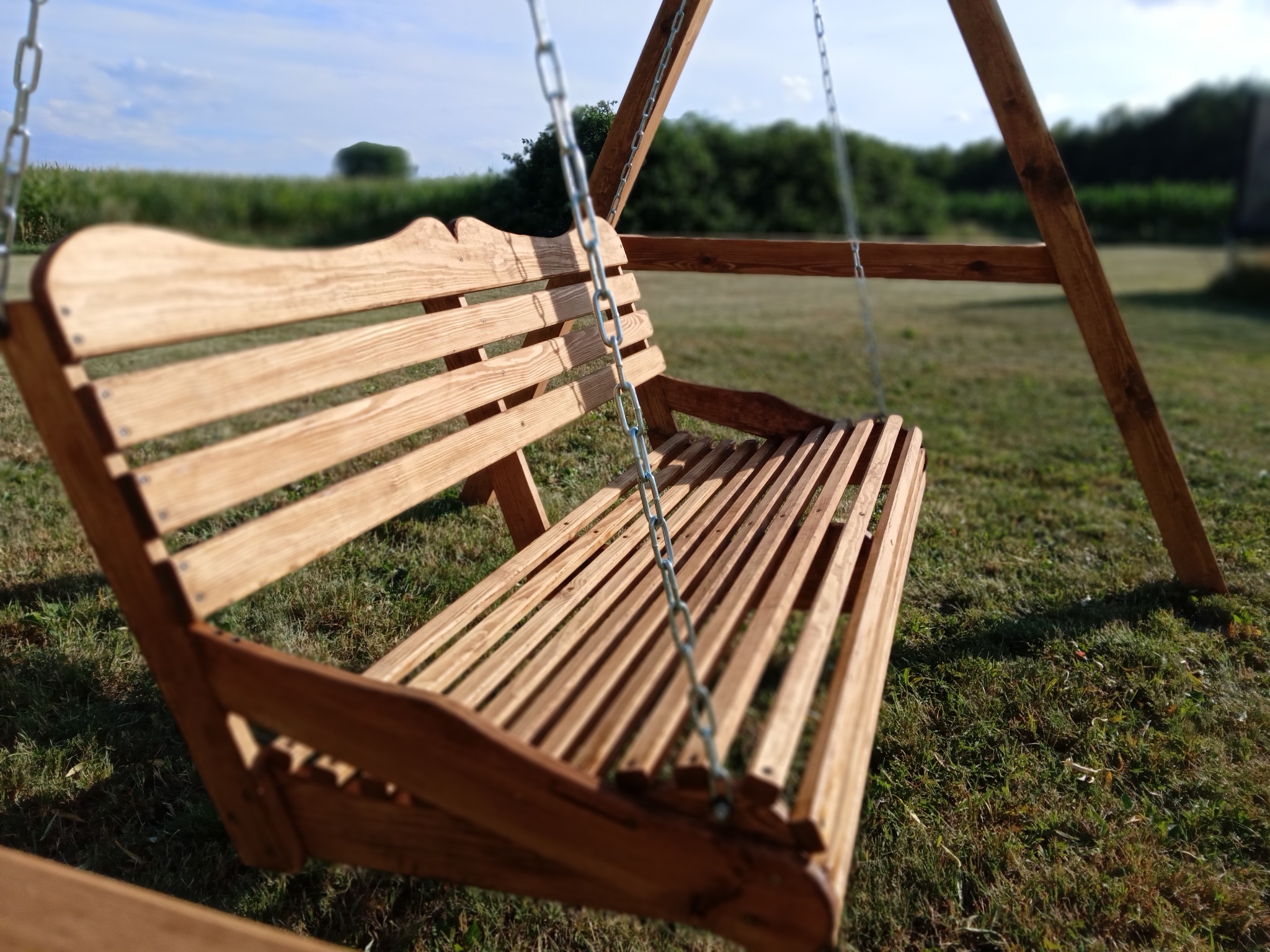 Drewniana huśtawka ogrodowa z oparciem o dekoracyjnym kształcie, zawieszona na łańcuchach. Tło to trawnik i zieleń, w oddali pole. Słoneczny dzień.