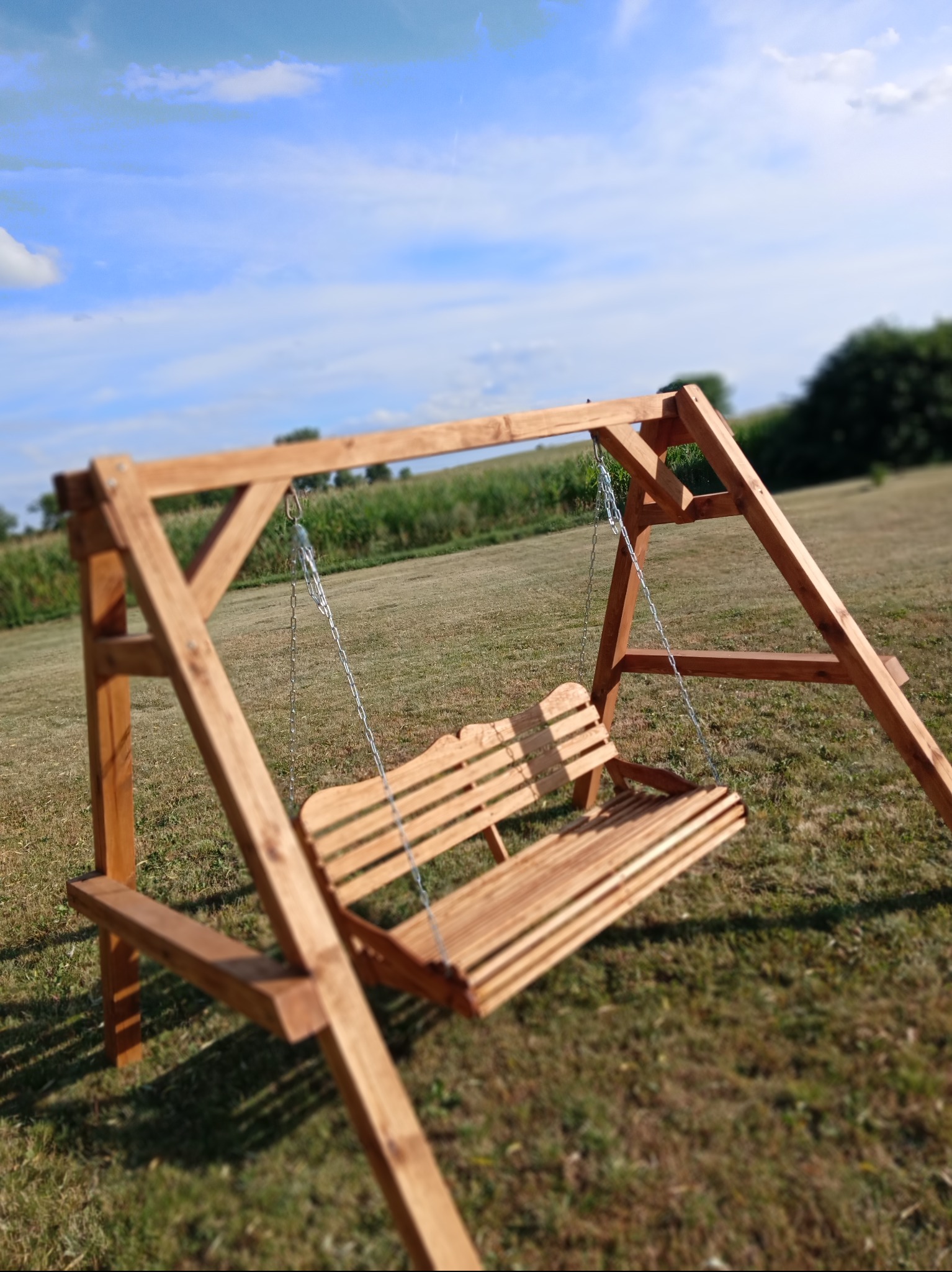 Drewniana huśtawka ogrodowa z oparciem, zawieszona na łańcuchach, ustawiona na trawniku. W tle łąka i błękitne niebo z chmurami.