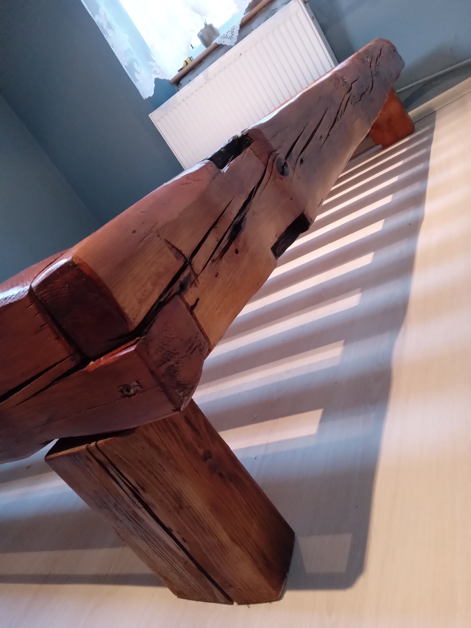 Fragment konstrukcji z surowego drewna o wyraźnych słojach, ustawiony ukośnie, rzucający cienie na ścianę. Widoczne detale obróbki drewna i naturalne pęknięcia.