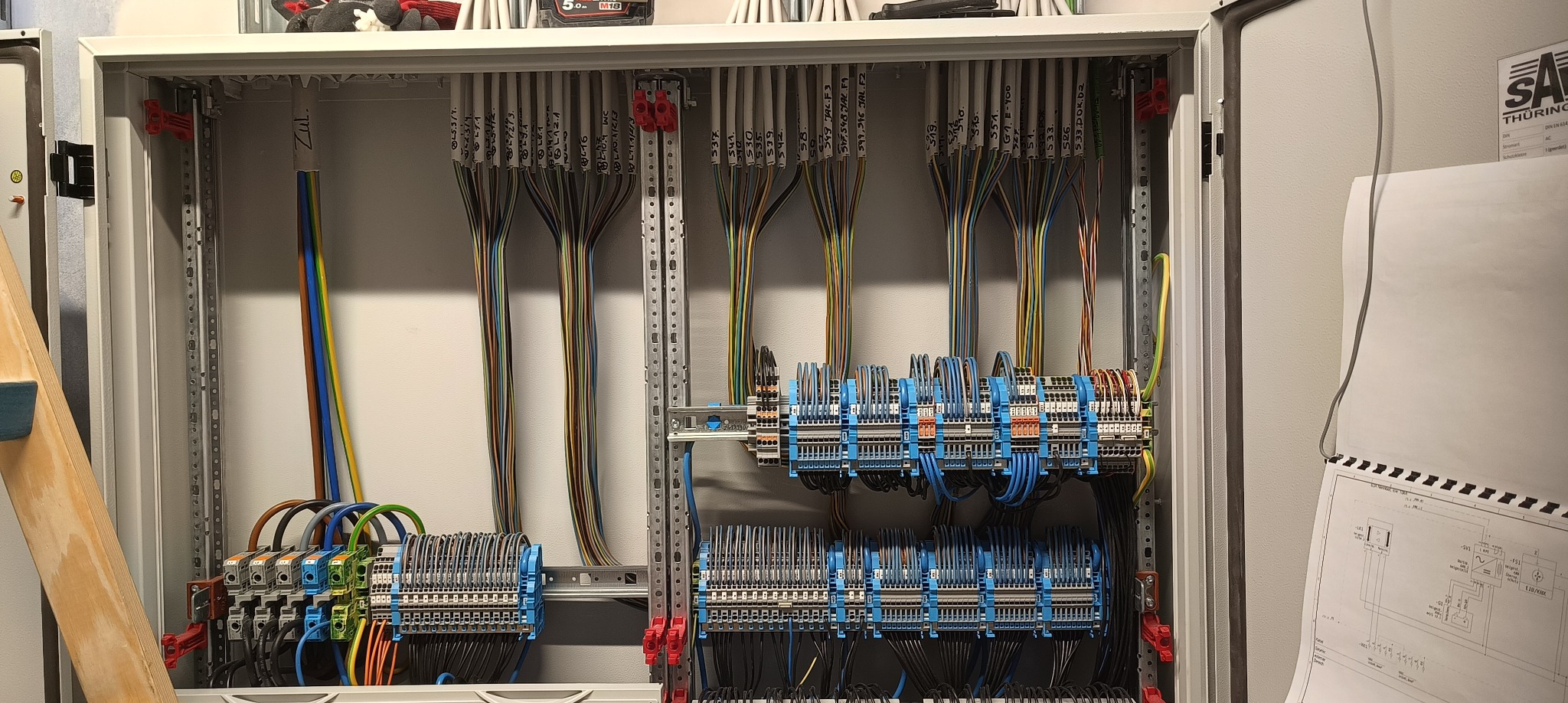 Szafa elektryczna z precyzyjnie ułożonymi przewodami, oznaczonymi etykietami. Widoczne listwy zaciskowe i schemat elektryczny po prawej stronie.