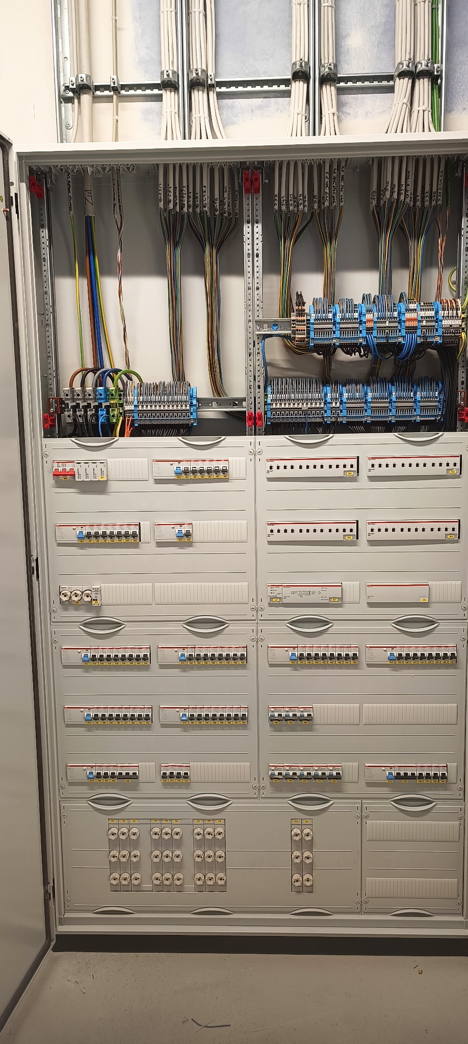 Rozdzielnica elektryczna w szarej obudowie z licznymi kablami oznaczonymi etykietami, widoczne bezpieczniki i elementy sterujące. Instalacja elektryczna wewnątrz budynku.