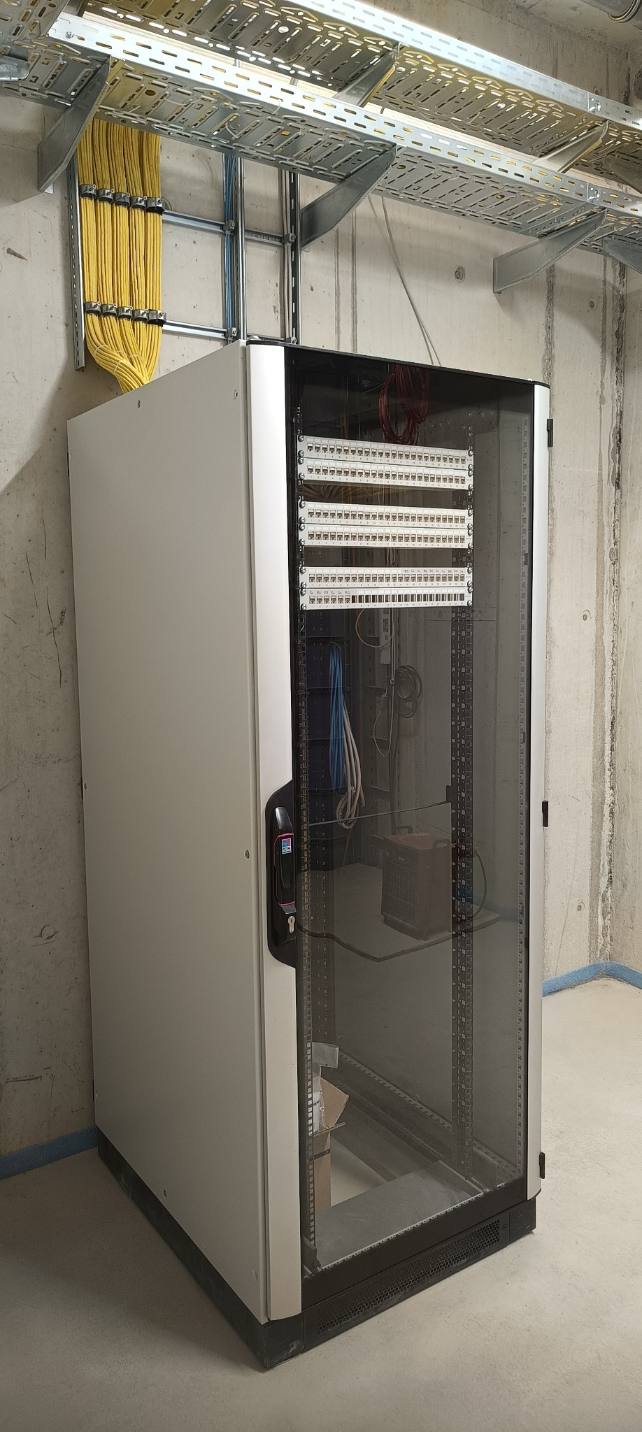 Szafa rack z panelami krosowymi i okablowaniem w surowym wnętrzu. Widoczne korytka kablowe pod sufitem i żółte przewody. Szara ściana z betonową fakturą.