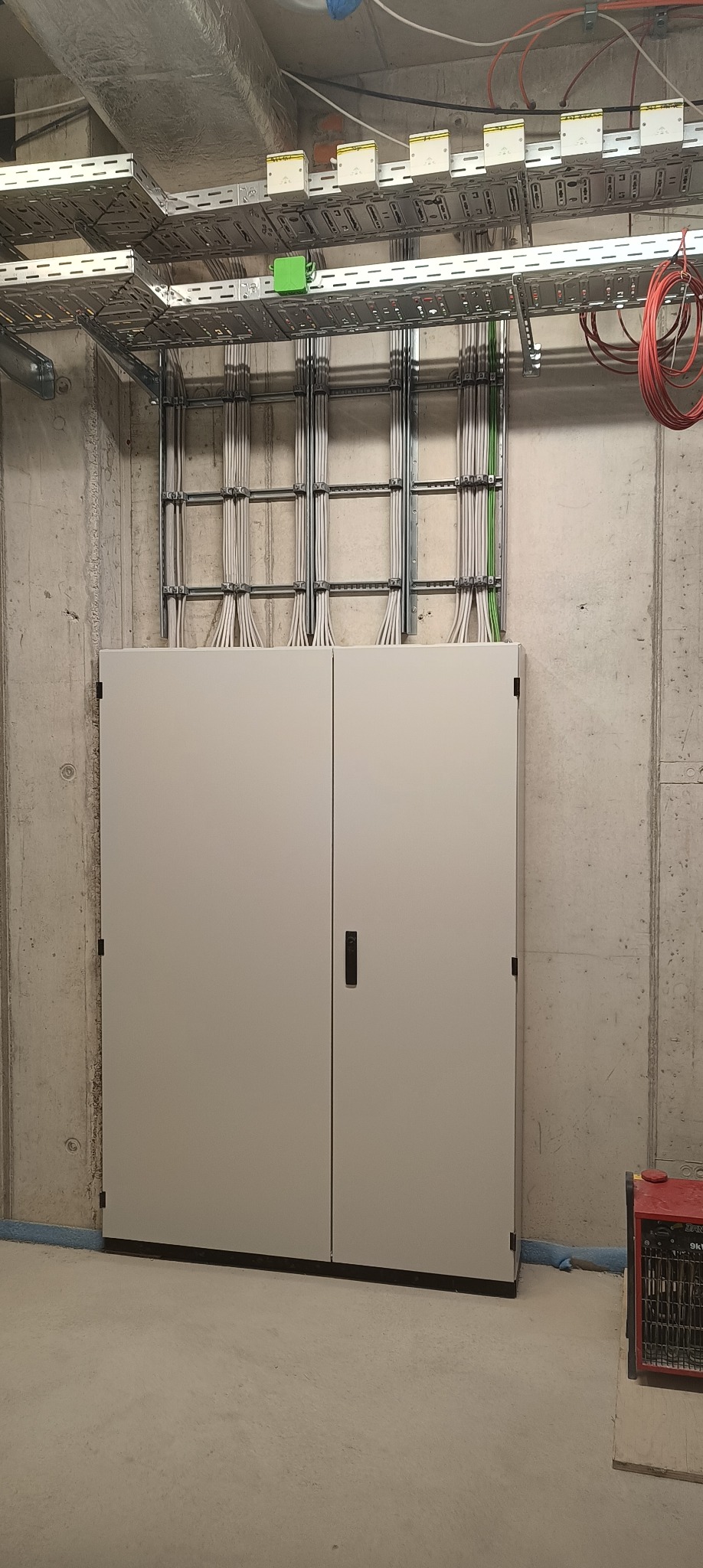 Szara szafa elektryczna z zamontowanymi kablami w metalowych korytach na betonowej ścianie. Widoczne kable w kolorze białym, zielonym i czerwonym.