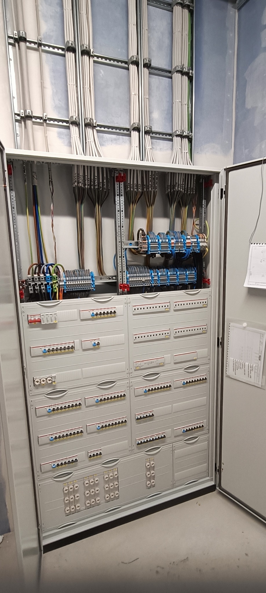 Otwarta szafa elektryczna z licznymi przewodami i bezpiecznikami, widoczne kable prowadzone w korytkach na ścianie. Instalacja w nowym budynku.