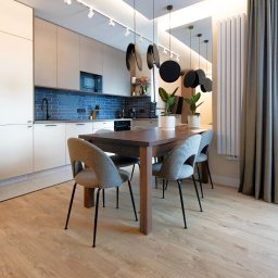 Gawra Studio Projektowe Justyna Miś-Nalepka - Nowoczesna kuchnia z jadalnią: drewniany stół otoczony krzesłami, minimalistyczne szafki, niebieskie płytki nad blatem i dekoracyjne lampy wiszące.