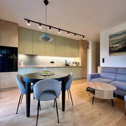 Gawra Studio Projektowe Justyna Miś-Nalepka - Jasne wnętrze: kuchnia z zielonymi frontami, okrągły stół z szarymi krzesłami, drewniana podłoga i szara sofa. Oświetlenie punktowe i obraz na ścianie.