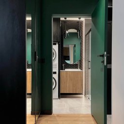 Gawra Studio Projektowe Justyna Miś-Nalepka - Nowoczesna łazienka z zielonymi ścianami, drewnianą podłogą i szafką pod umywalką. Widoczna pralka i suszarka w zabudowie. Oświetlenie LED podkreśla detale.