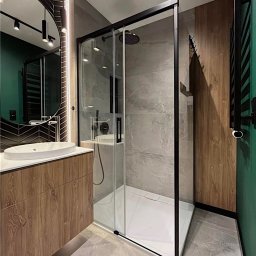 Gawra Studio Projektowe Justyna Miś-Nalepka - Nowoczesna łazienka z kabiną prysznicową typu walk-in, umywalką nablatową na drewnianej szafce i okrągłym lustrem z podświetleniem LED. Szare płytki i akcenty zieleni.