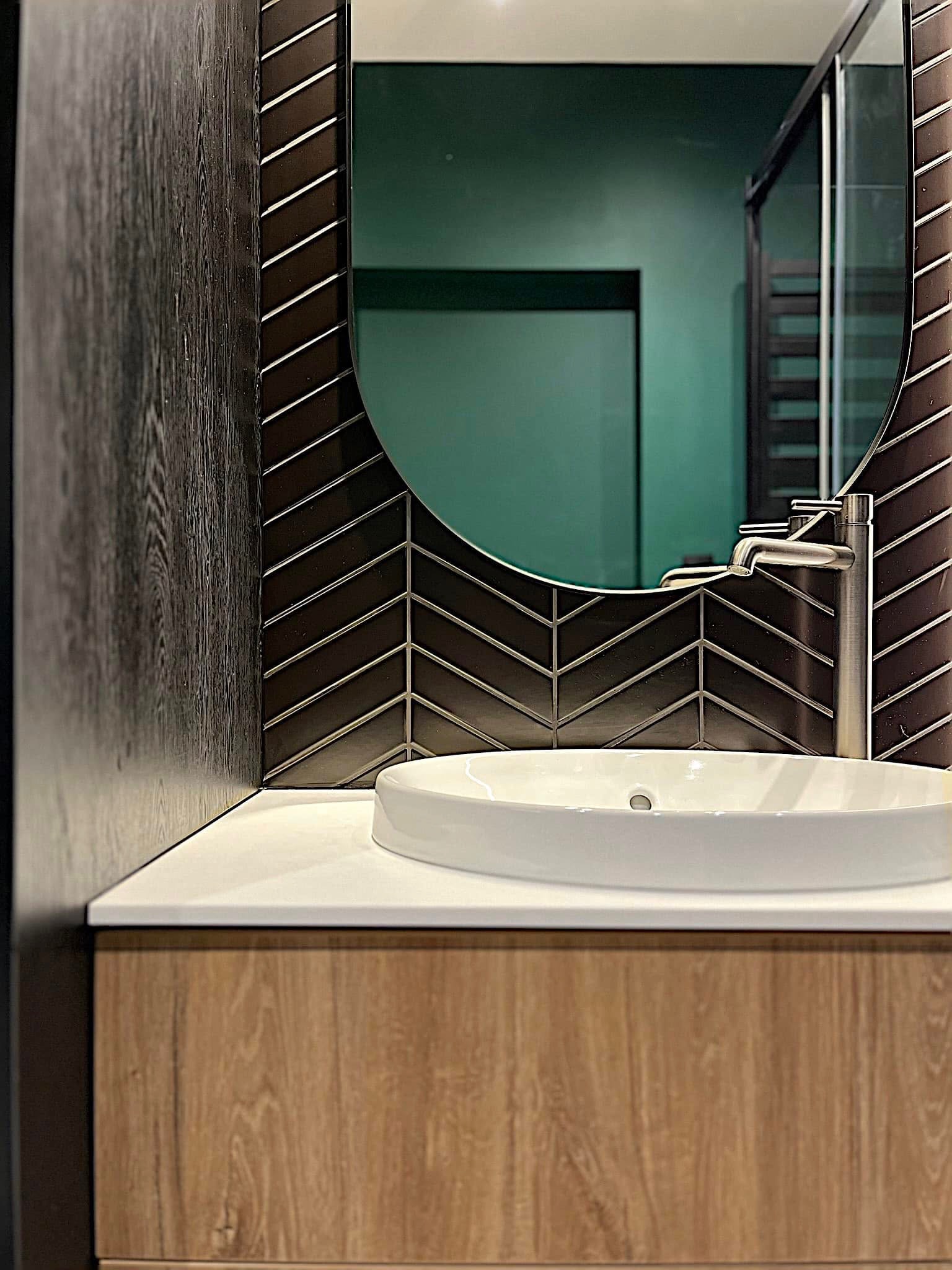 Nowoczesna umywalka z baterią na blacie z drewnianą szafką. W tle lustro w ramie z płytek ułożonych w jodełkę i ściana w odcieniach zieleni.
