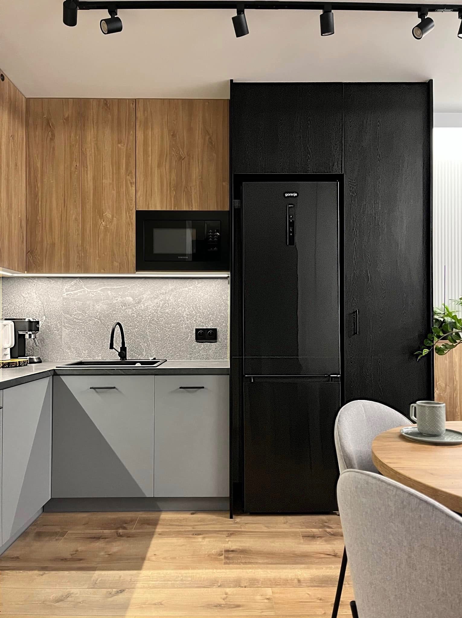 Nowoczesna kuchnia z drewnianymi i szarymi szafkami, czarnym sprzętem AGD (lodówka, mikrofalówka) i oświetleniem punktowym. Uwagę zwraca drewniana podłoga i okrągły stół.