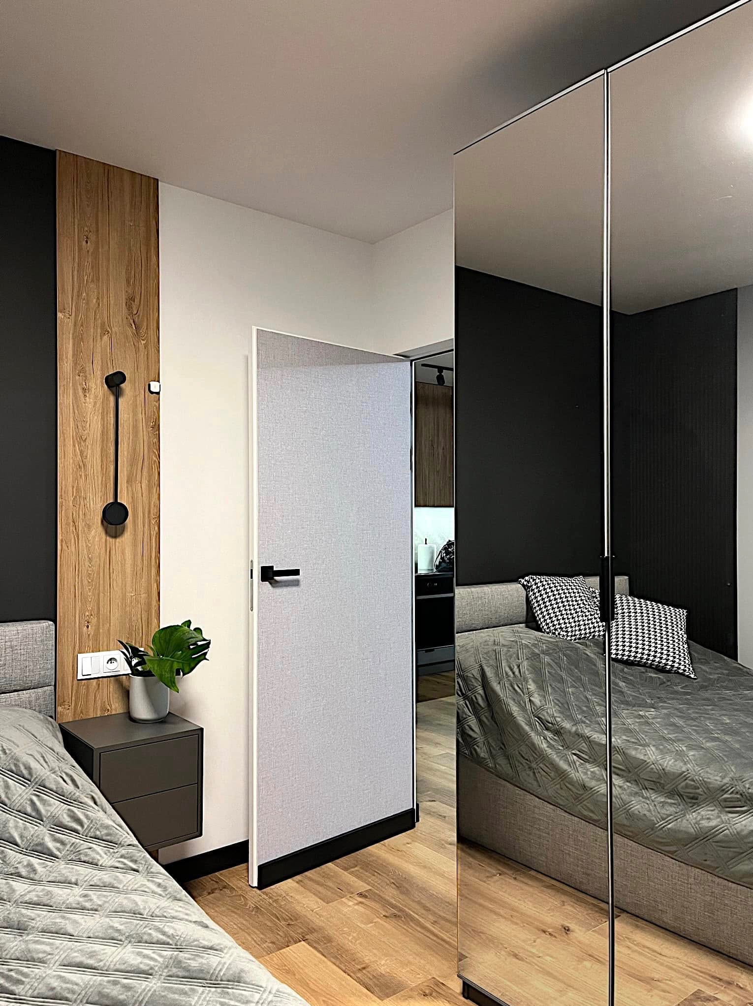 Nowoczesna sypialnia z łóżkiem, szafką nocną z rośliną i lustrzaną szafą. Kontrastowe połączenie czerni, bieli i drewna. Widoczne detale oświetlenia i wykończenia.