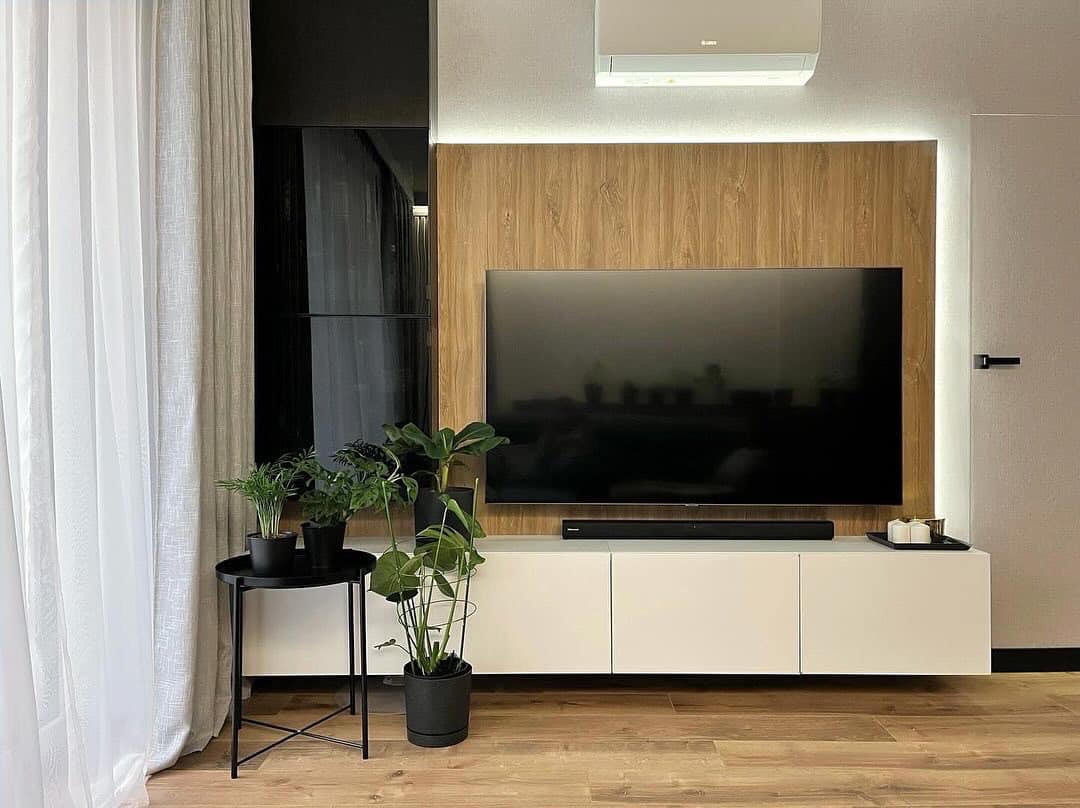 Nowoczesny salon z drewnianą ścianą, telewizorem, białymi szafkami i roślinami w doniczkach. Stonowana kolorystyka, minimalistyczny design i subtelne oświetlenie LED.
