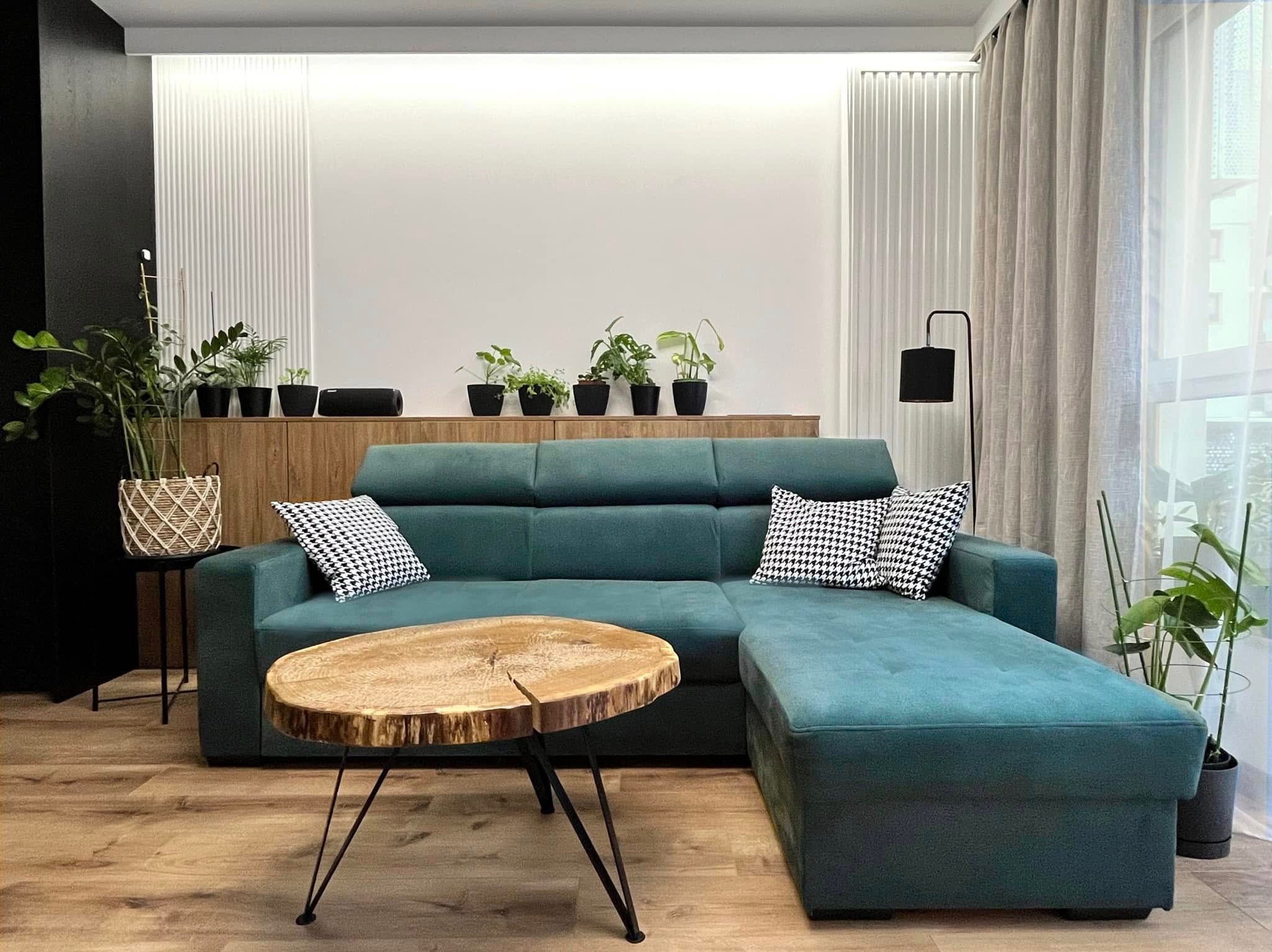 Turkusowa sofa narożna z drewnianym stolikiem kawowym na tle białej ściany z roślinami doniczkowymi. Nowoczesne wnętrze salonu z parkietem.