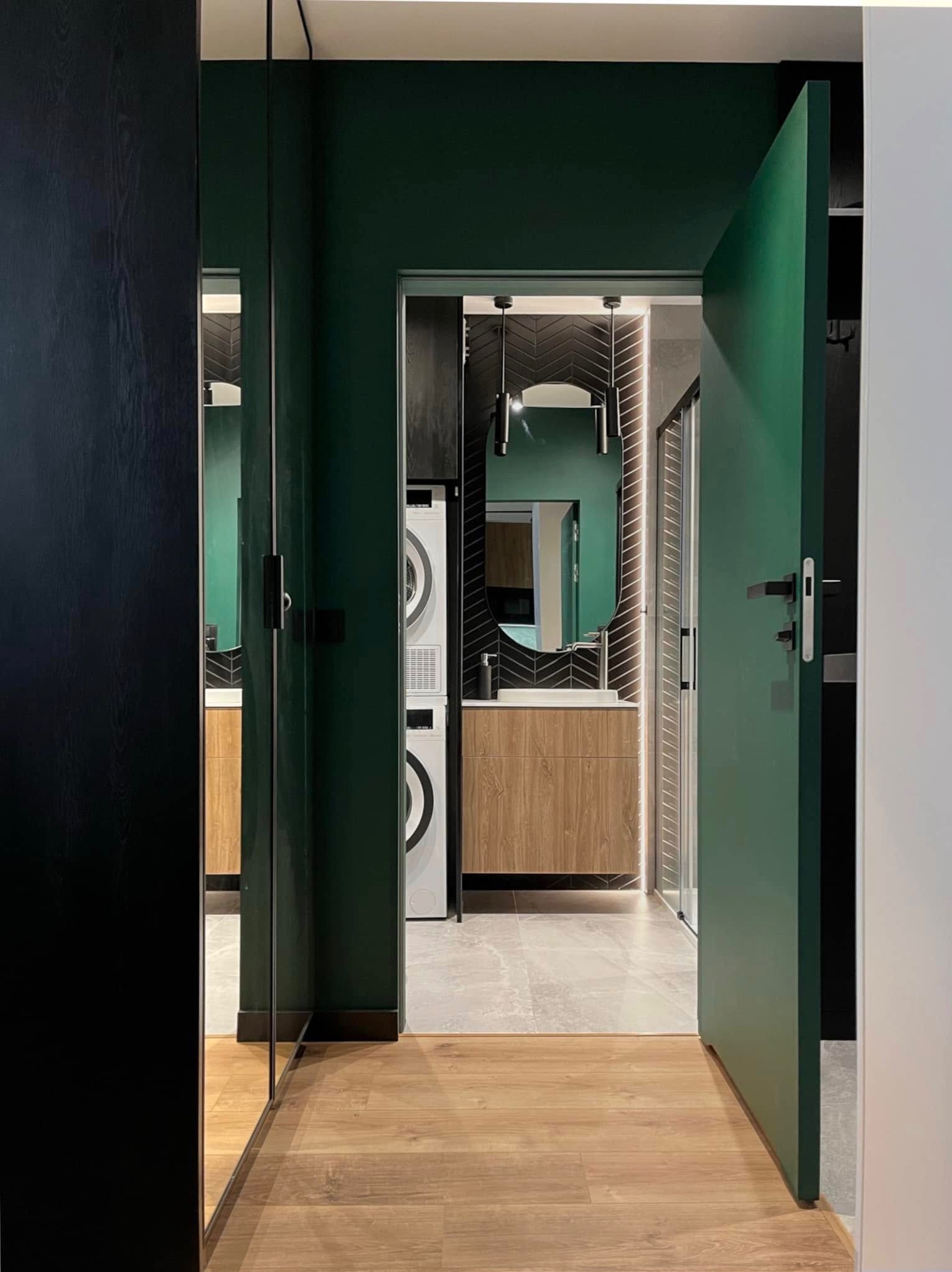 Nowoczesna łazienka z zielonymi ścianami, drewnianą podłogą i szafką pod umywalką. Widoczna pralka i suszarka w zabudowie. Oświetlenie LED podkreśla detale.