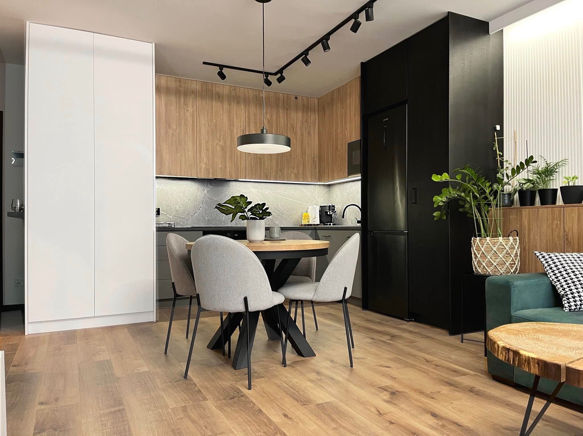 Nowoczesna kuchnia z okrągłym stołem i szarymi krzesłami. Drewniane fronty szafek, czarne akcenty, oświetlenie punktowe i rośliny doniczkowe tworzą przytulną atmosferę.