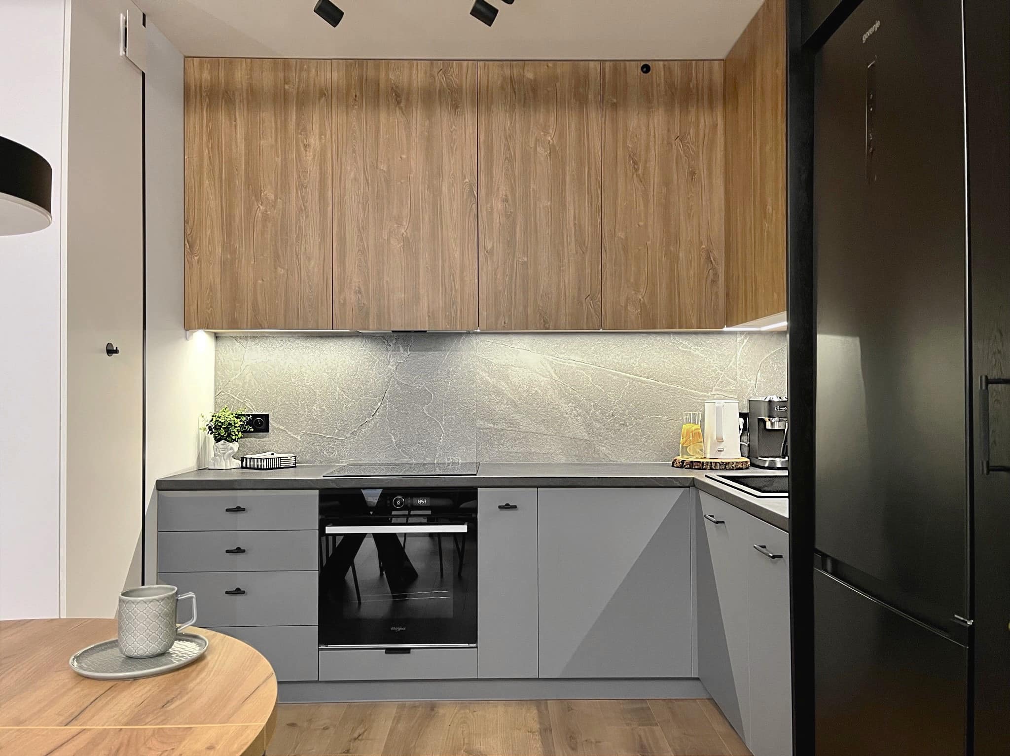 Nowoczesna kuchnia w szarościach z drewnianymi akcentami: szafki górne z forniru, szary blat i fronty, czarny piekarnik i lodówka. Minimalistyczny design.