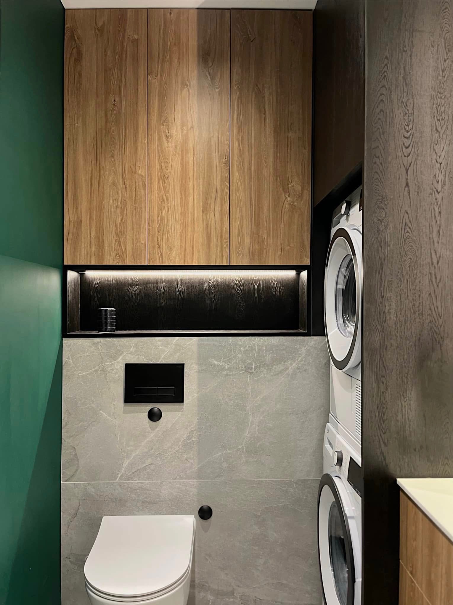 Nowoczesna łazienka z zabudową meblową z drewna, szarą płytką imitującą marmur i zieloną ścianą. Widoczna toaleta podwieszana i pralka z suszarką w zabudowie.