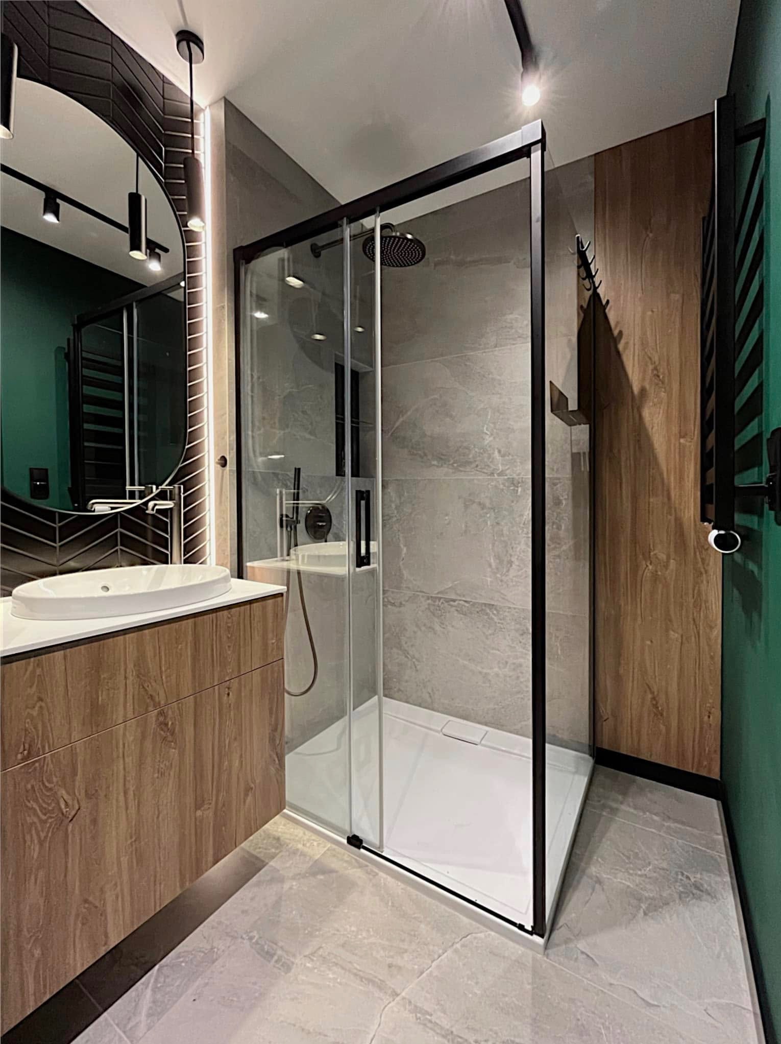 Nowoczesna łazienka z kabiną prysznicową typu walk-in, umywalką nablatową na drewnianej szafce i okrągłym lustrem z podświetleniem LED. Szare płytki i akcenty zieleni.