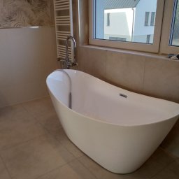 Mykola Kreshchyk - Wolnostojąca wanna z chromowaną baterią, grzejnik drabinkowy i okno z widokiem na dom w tle. Beżowe płytki na ścianach i podłodze, minimalistyczny wystrój.