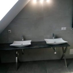 Mykola Kreshchyk - Nowoczesna łazienka na poddaszu: dwie białe, designerskie umywalki na blacie z ciemnego kamienia, podświetlane lampą, szare płytki na ścianach i podłodze.