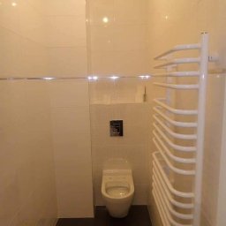 Mykola Kreshchyk - Wykończona łazienka w beżowych płytkach z podwieszaną toaletą i białym grzejnikiem drabinkowym. Ciemna podłoga i metalowy przycisk spłuczki.