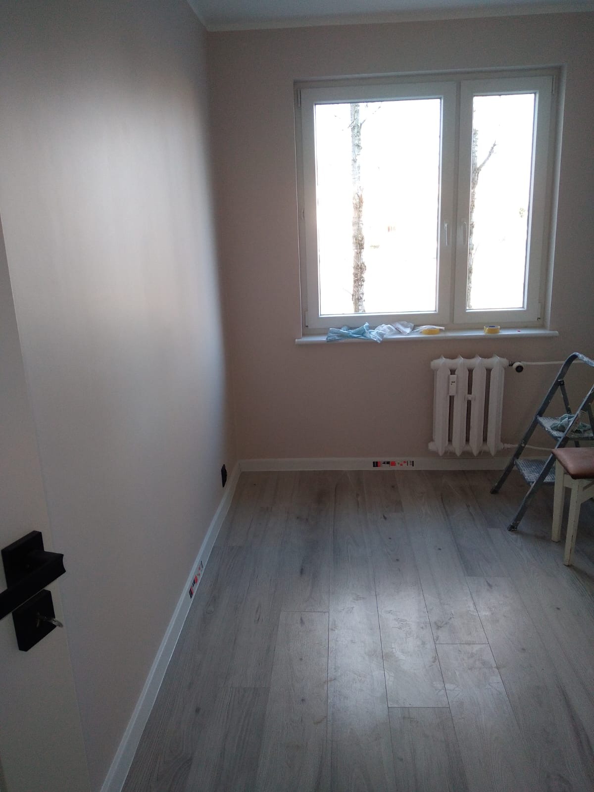 Jasne wnętrze po malowaniu ścian na beżowo, widoczne panele podłogowe, grzejnik, drabina malarska i okno. Świeżo wykończony pokój.