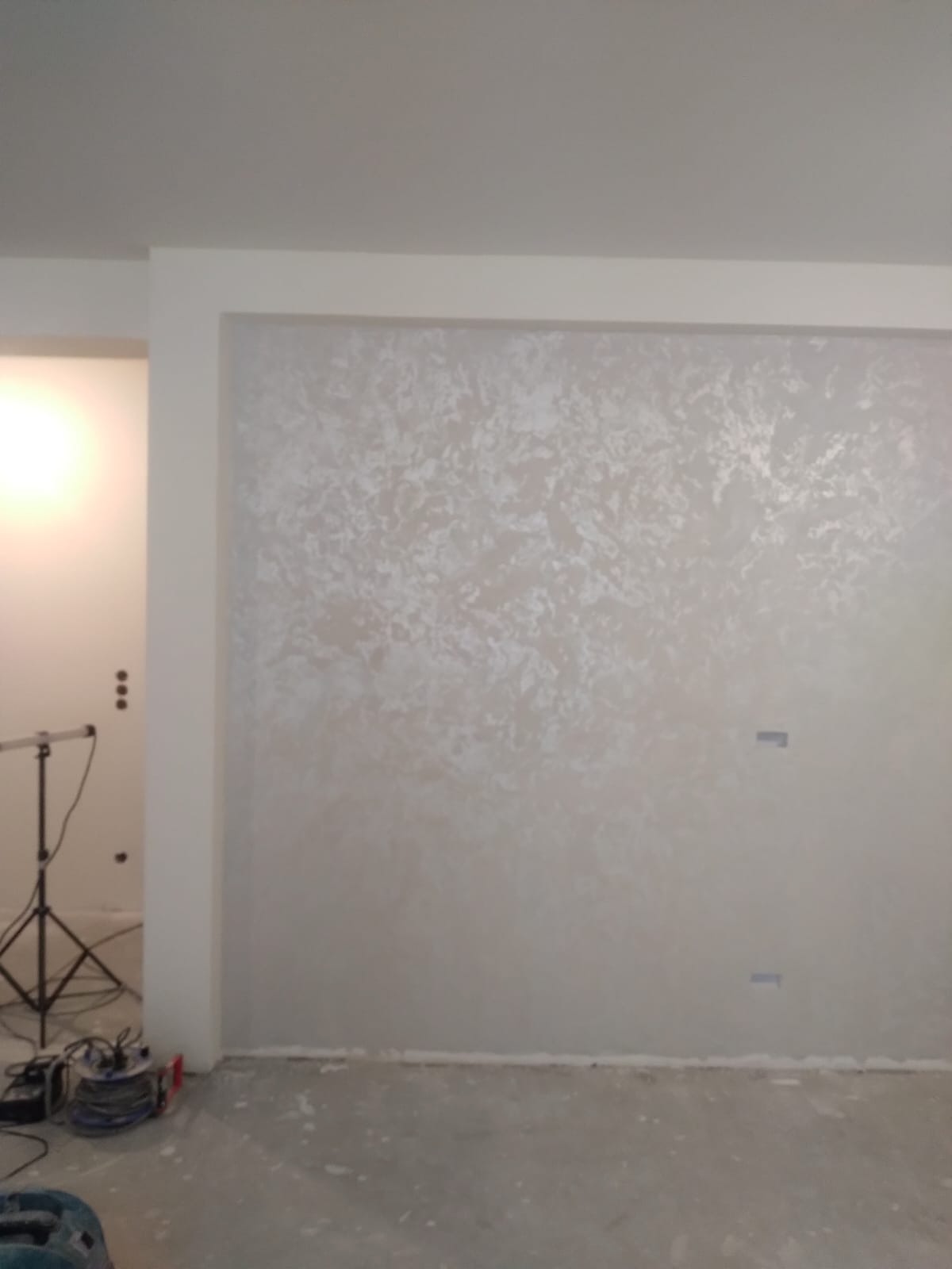 Ściana z efektem dekoracyjnym w odcieniach beżu i bieli, widoczna w pokoju w trakcie wykończenia, z lampą i narzędziami w tle.