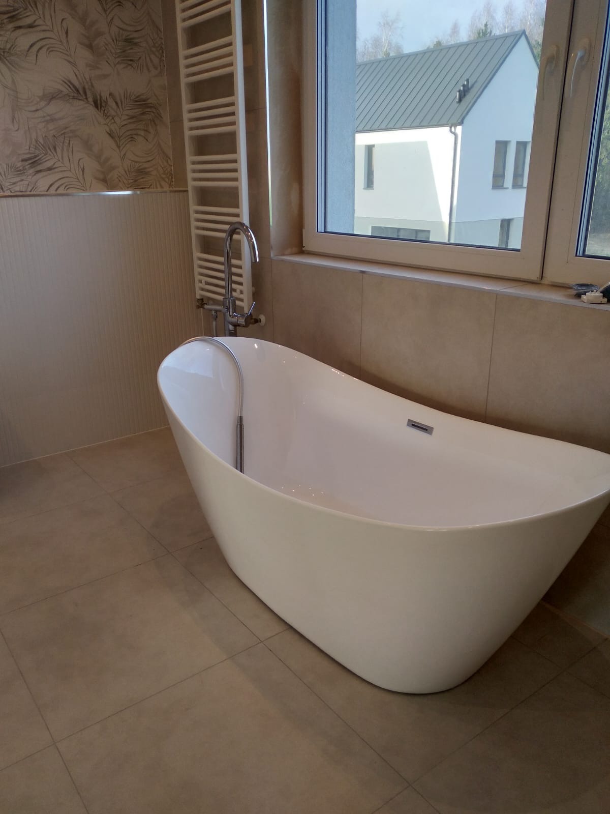 Wolnostojąca wanna z chromowaną baterią, grzejnik drabinkowy i okno z widokiem na dom w tle. Beżowe płytki na ścianach i podłodze, minimalistyczny wystrój.