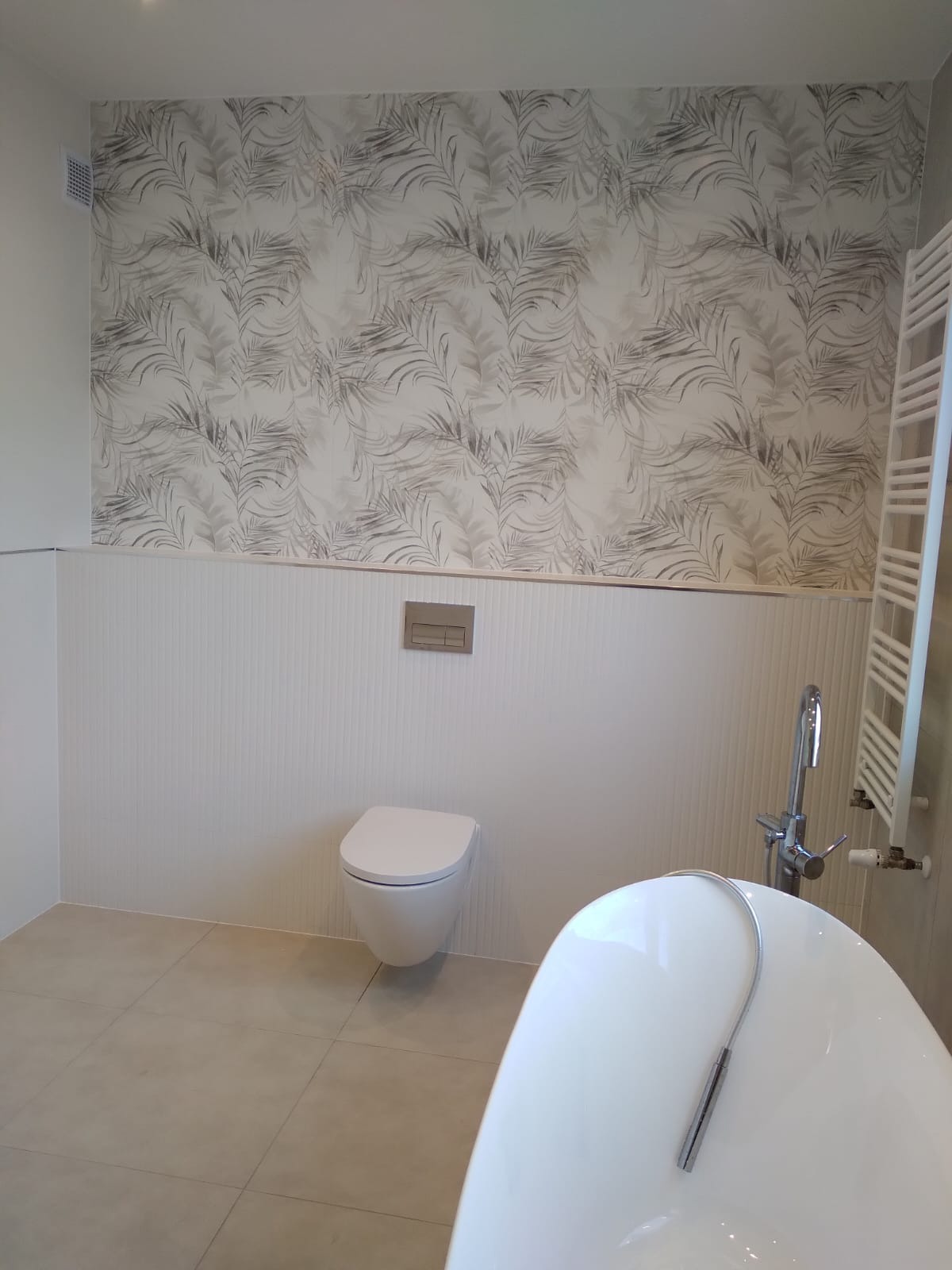 Elegancka łazienka: biała wanna, podwieszana toaleta, grzejnik drabinkowy i ściana z motywem liści. Nowoczesny design i jasna kolorystyka.