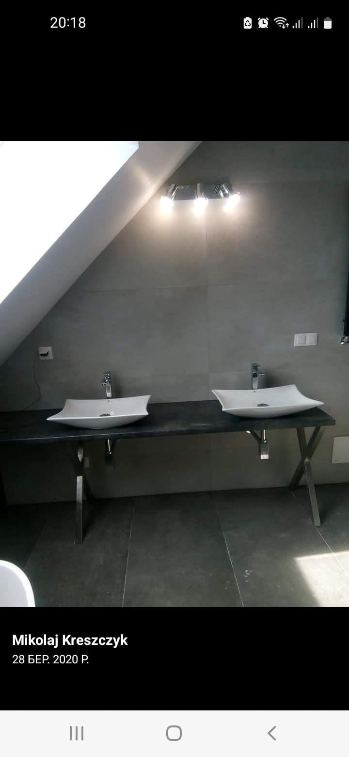 Nowoczesna łazienka na poddaszu: dwie białe, designerskie umywalki na blacie z ciemnego kamienia, podświetlane lampą, szare płytki na ścianach i podłodze.