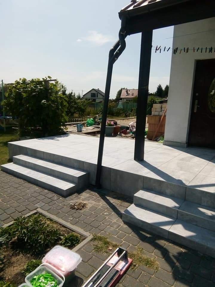 Wykończony taras z szarych płyt, widoczne schody wejściowe. Na pierwszym planie kostka brukowa i narzędzia. W tle dom z podwieszonymi klamerkami.