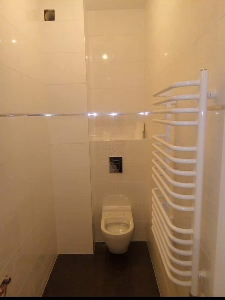 Wykończona łazienka w beżowych płytkach z podwieszaną toaletą i białym grzejnikiem drabinkowym. Ciemna podłoga i metalowy przycisk spłuczki.