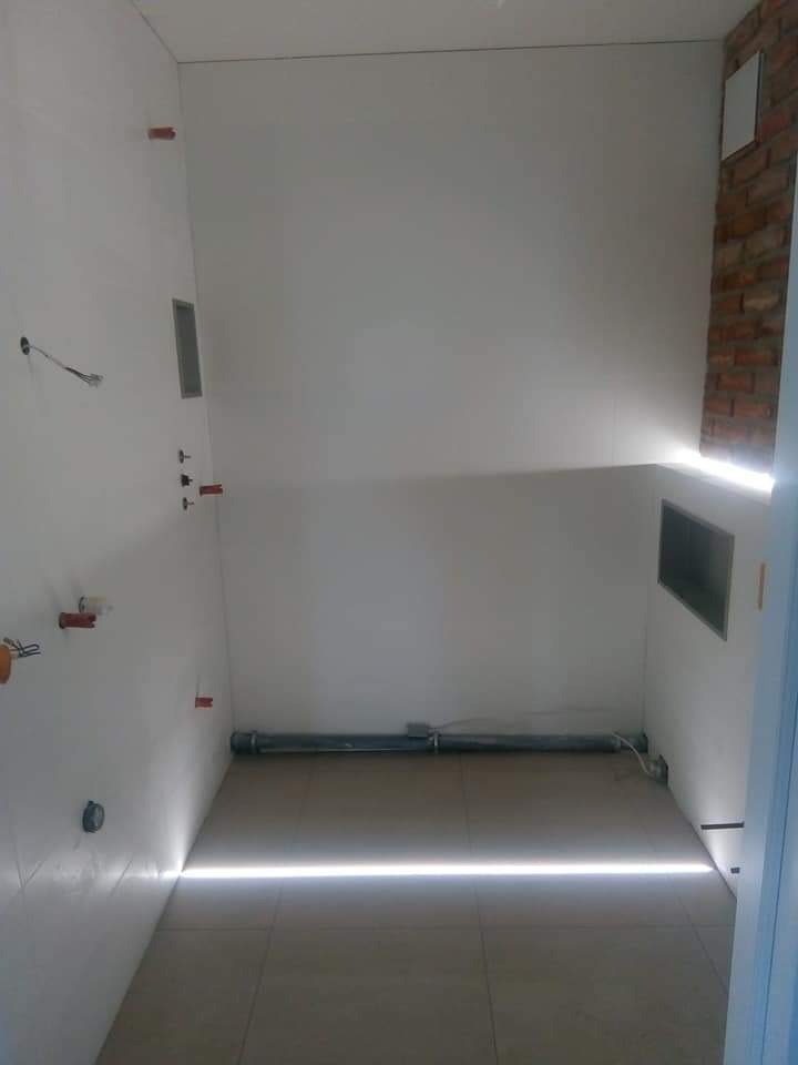 Wnętrze łazienki w trakcie prac wykończeniowych: białe płytki ścienne, fragment ściany z cegły, widoczne instalacje i rura odpływowa wzdłuż podłogi z płytek.