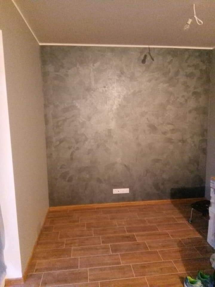 Pokój po remoncie: drewnopodobna podłoga z płytek, ściana z efektem dekoracyjnym w odcieniach szarości. Widoczne gniazdko i niedokończone oświetlenie sufitowe.