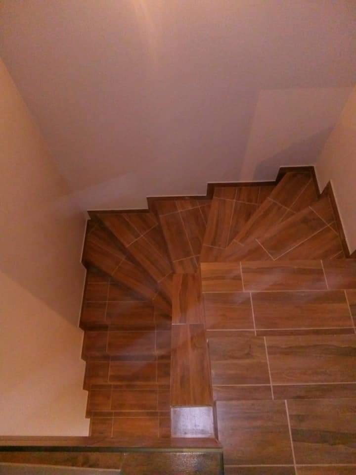 Wykończone schody z brązowych płytek imitujących drewno, widok z góry. Stopnie ułożone spiralnie, jasne ściany i sufit. Widoczny fragment dolnego stopnia.