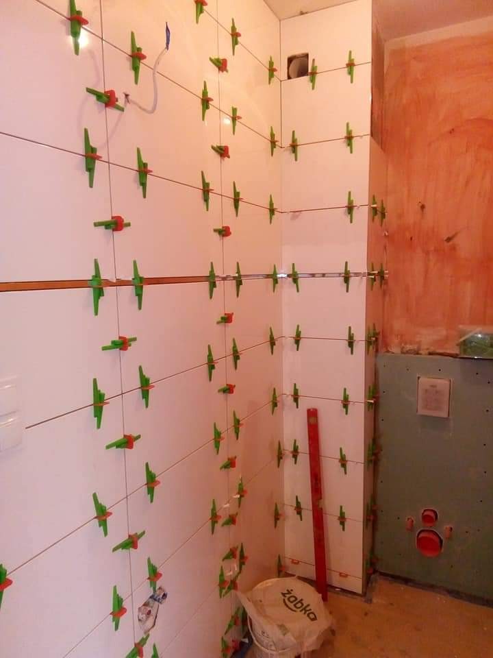 Ściana z białych płytek, w trakcie układania, z systemem poziomowania (zielone krzyżyki i czerwone kliny). Widoczny poziomica i wiadro po kleju.