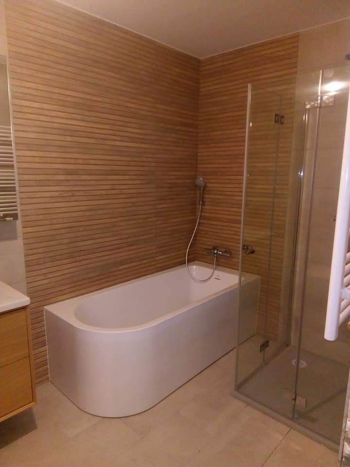 Nowoczesna łazienka z drewnopodobną okładziną ścienną, białą wanną i kabiną prysznicową ze szkła. Minimalistyczny design i stonowana kolorystyka.