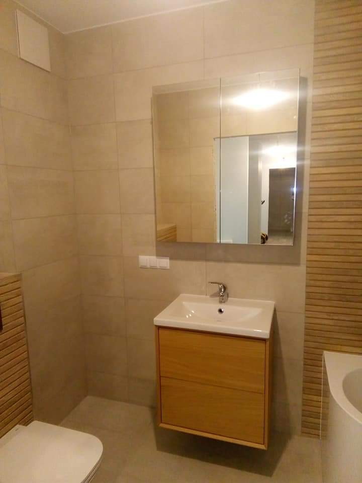 Wykończona łazienka w beżowej tonacji: płytki, lustro z szafką, umywalka z drewnianą szafką. Minimalistyczny design, jasne wnętrze. Widoczne detale wykończenia.