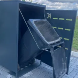 Metalowa obudowa śmietnika w kolorze grafitowym, z ażurowym wzorem i roślinami na dachu, umieszczona na brukowanej powierzchni.