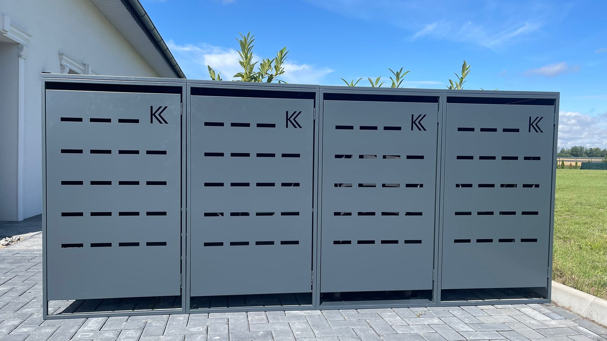 Cztery szare, metalowe konstrukcje z wyciętymi poziomymi paskami i literą 'K' na tle błękitnego nieba i zieleni, stojące na szarej kostce brukowej.