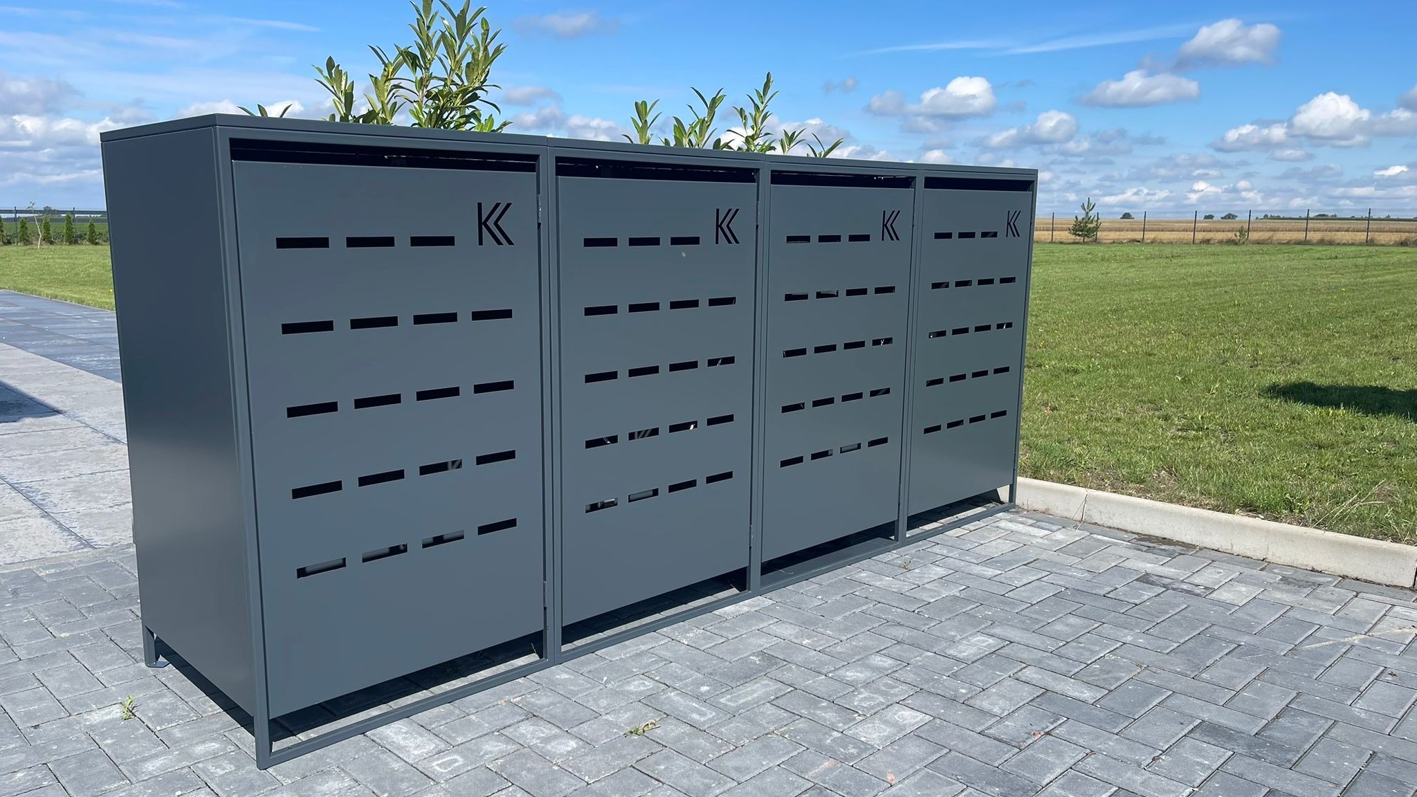 Cztery szare, metalowe szafki na rośliny z wyciętymi poziomymi szczelinami i logo 'K', ustawione na brukowanym podłożu, z zielenią w tle i błękitnym niebem.