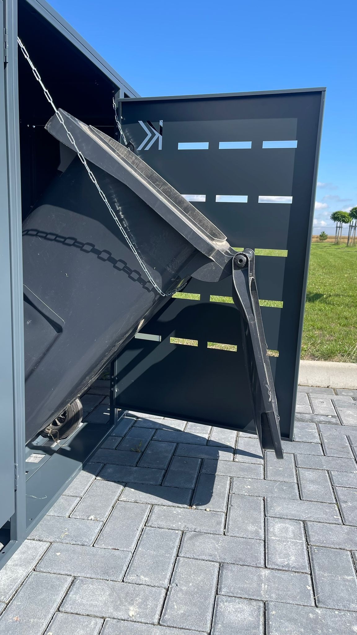 Metalowa obudowa na pojemniki na śmieci w kolorze antracytowym, z otwartymi drzwiami i podniesionym pojemnikiem, na tle kostki brukowej i zieleni.